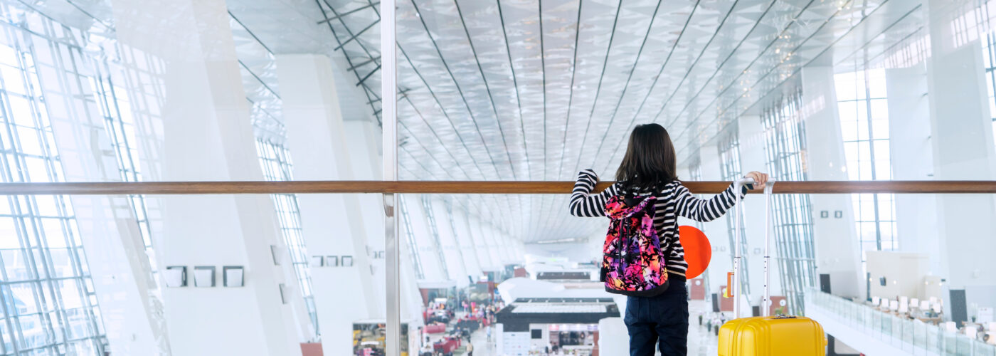 Child overlooking an airport terminal from above next to a yellow rolling suitcase