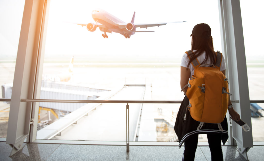 Women wearing yellow backpack and looking out of airport terminal window at an airplane