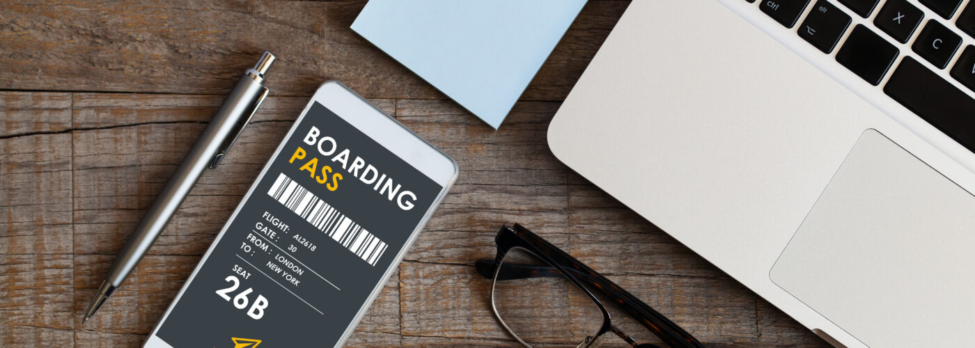 Phone showing digital boarding pass on a table next to a laptop, glasses, pen, and memo note pad