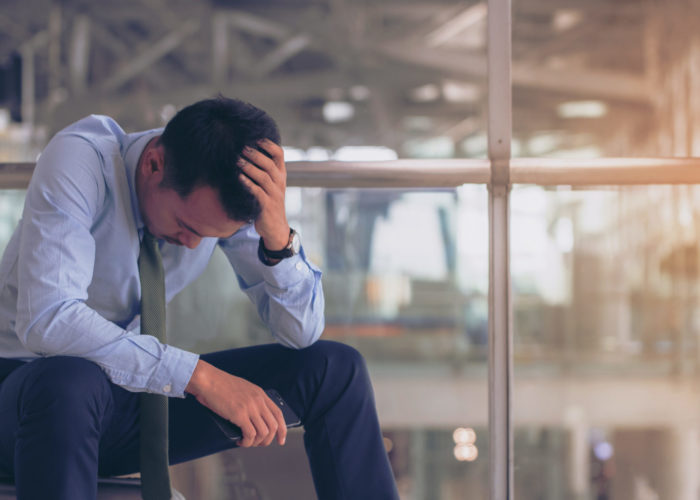 Man sitting on rolling luggage with his head in his hands on the upper level of an airport with a terminal in the background behind a glass window