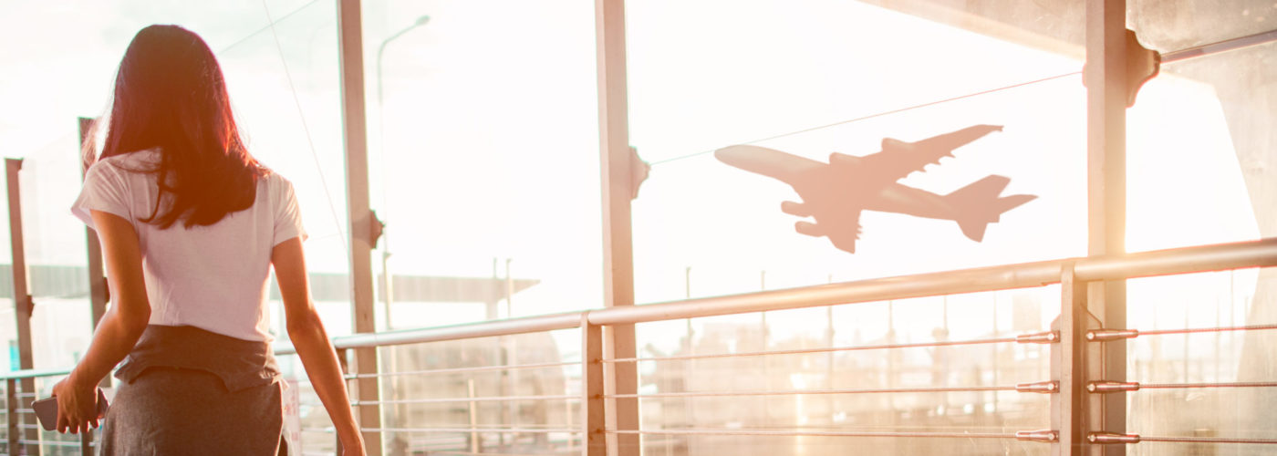 Woman pulling suitcase down hallway in airport next to glass window, out of which is a view of a plane taking off