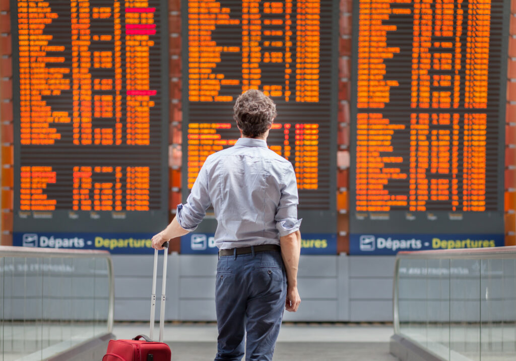 Man waiting with rolling luggage, looking up at departure board at an airport