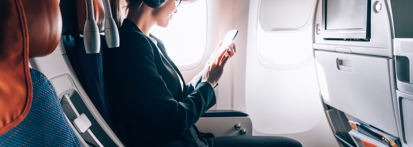 Woman sitting in window seat on airplane listening to media on her phone using noise cancelling headphones