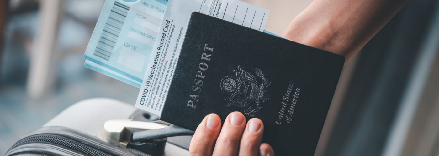 A person holding their COVID-19 vaccine card, passport, boarding pass, and suitcase