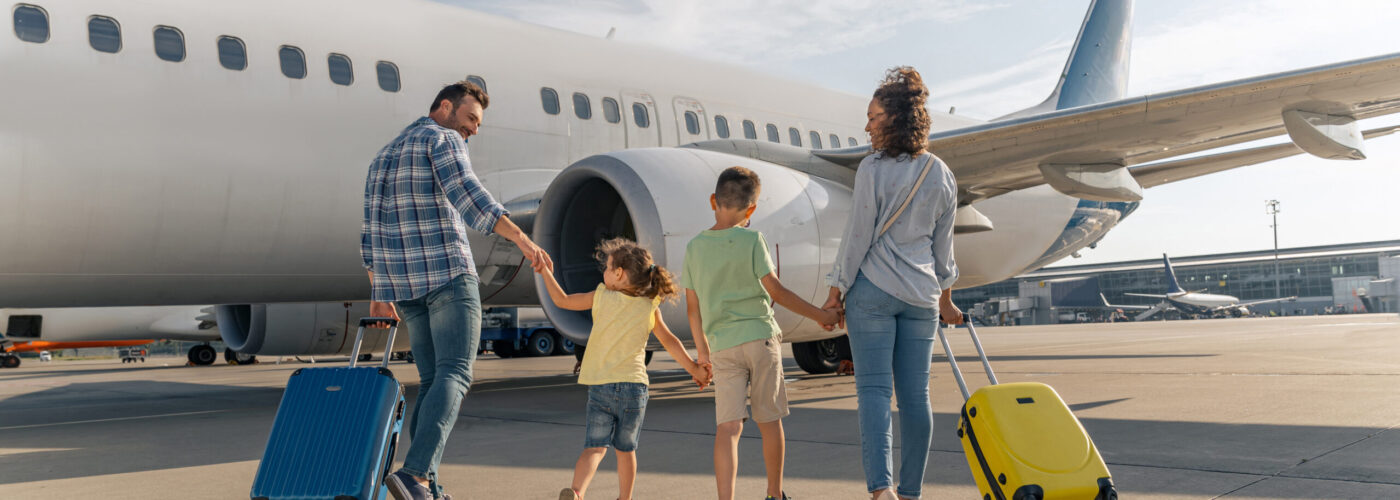 Parents and children walking toward plane on tarmac while wheeling suitcases