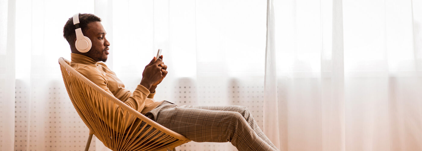 Man In Wireless Headset Using Mobile Phone Listening To Audiobook Sitting On Modern Chair Against Window Indoor