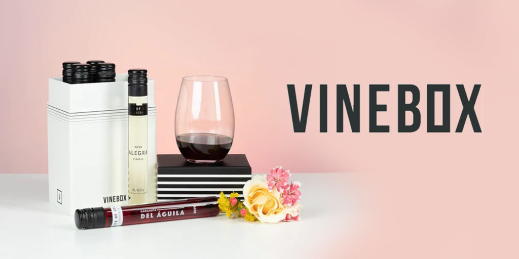 Four vials of wine in a white box with two vials placed outside the box next to a partially full wineglass, on a white table with a pink background. The VINEBOX logo is next to it.